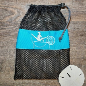 May include: Black mesh drawstring bag with a turquoise band featuring a white logo of a gymnast. The bag has a gray drawstring and a black toggle. A white sand dollar is in the lower right corner.
