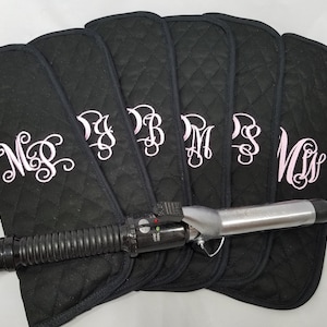 May include: Black quilted curling iron travel cases with embroidered initials in a light pink script. A black curling iron is placed on top of the cases. The cases are designed to protect the curling iron during travel.