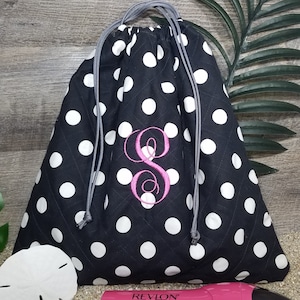 May include: Black drawstring bag with white polka dots and a pink embroidered letter "S". The bag has a gray drawstring closure and a quilted pattern. A pink Revlon hair dryer is visible in the foreground.