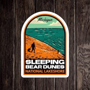 May include: A white sticker with a blue, green, and orange illustration of a beach scene with two people walking on a sand dune. The text "Michigan", "Sleeping Bear Dunes National Lakeshore" is printed on the sticker.
