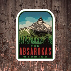 May include: A vintage-style travel sticker featuring a mountain range with a peak in the background. The sticker has a white border and says "THE ABSAROKAS WYOMING" in red and black text.