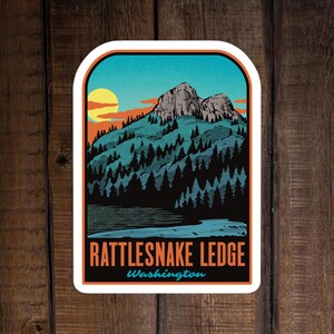 May include: A sticker featuring a mountain landscape with a lake and forest. The design includes a yellow sun, orange clouds, and the text "Rattlesnake Ledge Washington" in orange and blue. The sticker has a white border and is set against a wood grain background.