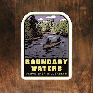 May include: A sticker with a vintage illustration of two people canoeing on a lake in a forest. The sticker has the text "Boundary Waters Canoe Area Wilderness" in yellow letters on a black background.