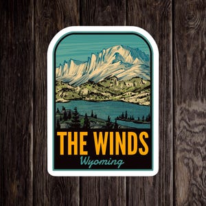 Puede incluir: Una pegatina de estilo vintage con una vista panorámica de The Winds en Wyoming. La pegatina representa una cordillera con picos cubiertos de nieve, un lago y árboles en tonos verdes y azules. El texto "THE WINDS" y "Wyoming" se muestra en amarillo.