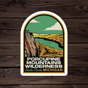 May include: A vintage-style sticker featuring a scenic view of a river winding through a forest with fall foliage. The sticker reads "Porcupine Mountains Wilderness State Park Michigan".