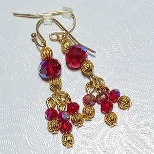 May include: Gold dangle earrings with red crystal beads. The earrings feature a larger red crystal bead at the top and smaller red crystal beads dangling below.