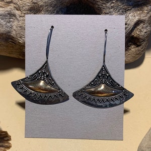 May include: A pair of silver-toned earrings with a fan-shaped design. The earrings feature a central gold-colored section and are adorned with intricate detailing and small, round embellishments. The earrings hang from curved silver hooks.