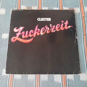 May include: A black vinyl record album cover with the title "Zuckerzeit" in pink and red lettering. The band name "CLUSTER" is printed in white above the title.