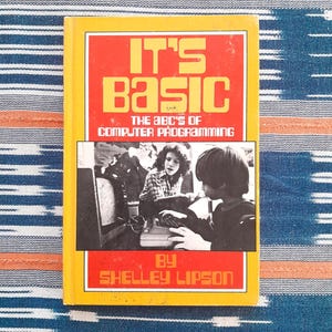 May include: A vintage book titled "IT'S BASIC" with the subtitle "The ABC's of Computer Programming" by Shelley Lipson. The cover is yellow with red and white text and a black and white photo of two people using a computer. The book is on a blue and white patterned surface.
