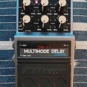May include: A Maxon DM-01 Multimode Delay digital effects pedal. The pedal has a black base with six circular indentations and a silver top with six knobs labeled "Min" and "Max". The pedal has a red "Effect" light and a black power cable.