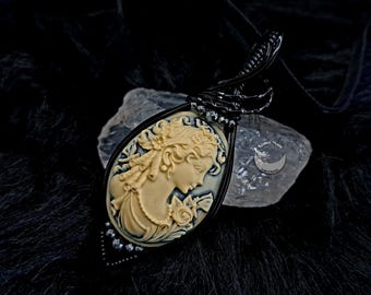 May include: A black wire-wrapped pendant featuring a cameo of a woman's profile in a white cameo. The cameo is set in a black metal frame and has a black wire wrapped around it. The pendant is hanging from a black cord.
