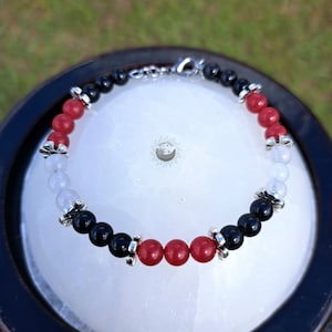 May include: A bracelet with black, red, and white beads, accented with silver charms. The bracelet is made with natural stones and is a stylish accessory.