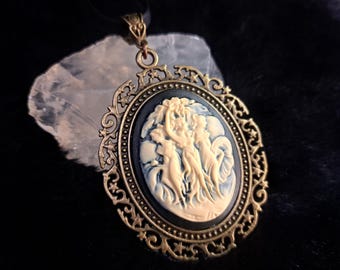 May include: An oval cameo pendant featuring a cream-colored relief of three figures against a black background, set within an ornate antique brass frame with star cutouts. The pendant hangs from a black cord and is positioned over a clear crystal.