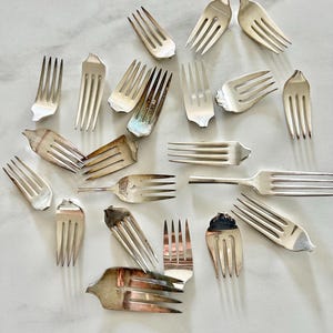 May include: A collection of silver forks, some with tines bent or broken, arranged in a circular pattern on a white marble surface.