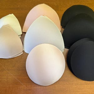 May include: A collection of bra inserts in various colors, including white, beige, pink, and black. The inserts are in different shapes, including triangular and rounded, and are made of a soft, padded material.