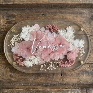 May include: An oval clear acrylic sign featuring the name "Vanessa" in white script, decorated with pink and white dried flowers. The sign is fixed with gold screws. The background is a wooden surface with a rustic appearance.