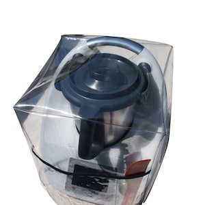 Puede incluir: Una cubierta de plástico transparente para un electrodoméstico de cocina. El electrodoméstico es un procesador de alimentos plateado con una tapa negra y un asa plateada. La cubierta tiene una cremallera negra y una espátula roja es visible en el interior.