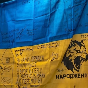 May include: A Ukrainian flag with blue and yellow sections, covered in handwritten messages and drawings. The flag features a cartoon character, a wolf illustration, and phrases like "Together to Victory" and "With us is truth."