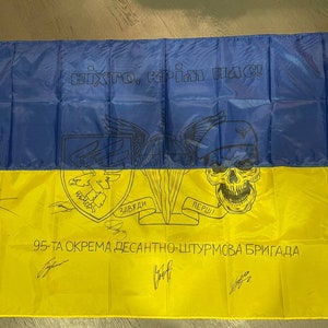Può includere: Una bandiera ucraina gialla e blu con un design di teschio e ossa incrociate. La bandiera ha il testo "Ніхто, крім нас!" e "95-ТА ОКРЕМА ДЕСАНТНО-ШТУРМОВА БРИГАДА" stampato su di essa.