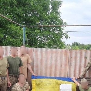 May include: A group of people in military uniforms stand in front of a yellow and blue flag with handwritten messages. The flag is draped over a table and the people are smiling.