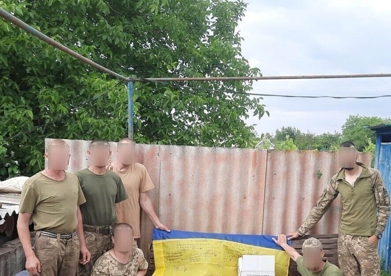 May include: A group of people in military uniforms stand in front of a yellow and blue flag with handwritten messages. The flag is draped over a table and the people are smiling.