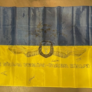 May include: A yellow and blue Ukrainian flag with a drawing of a tank and the text "Glory to the heroes! We are the glory of Ukraine - we will be free!"
