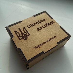 May include: A small wooden box with a brown and black finish. The box has a Ukrainian coat of arms and the text "Ukraine Artifact" on the top. The bottom of the box has the text "Борись-за-свободу" in Cyrillic.