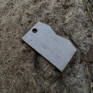 May include: A silver metal tag with a rounded edge and a hole in the top. The tag is lying on a bed of sand.