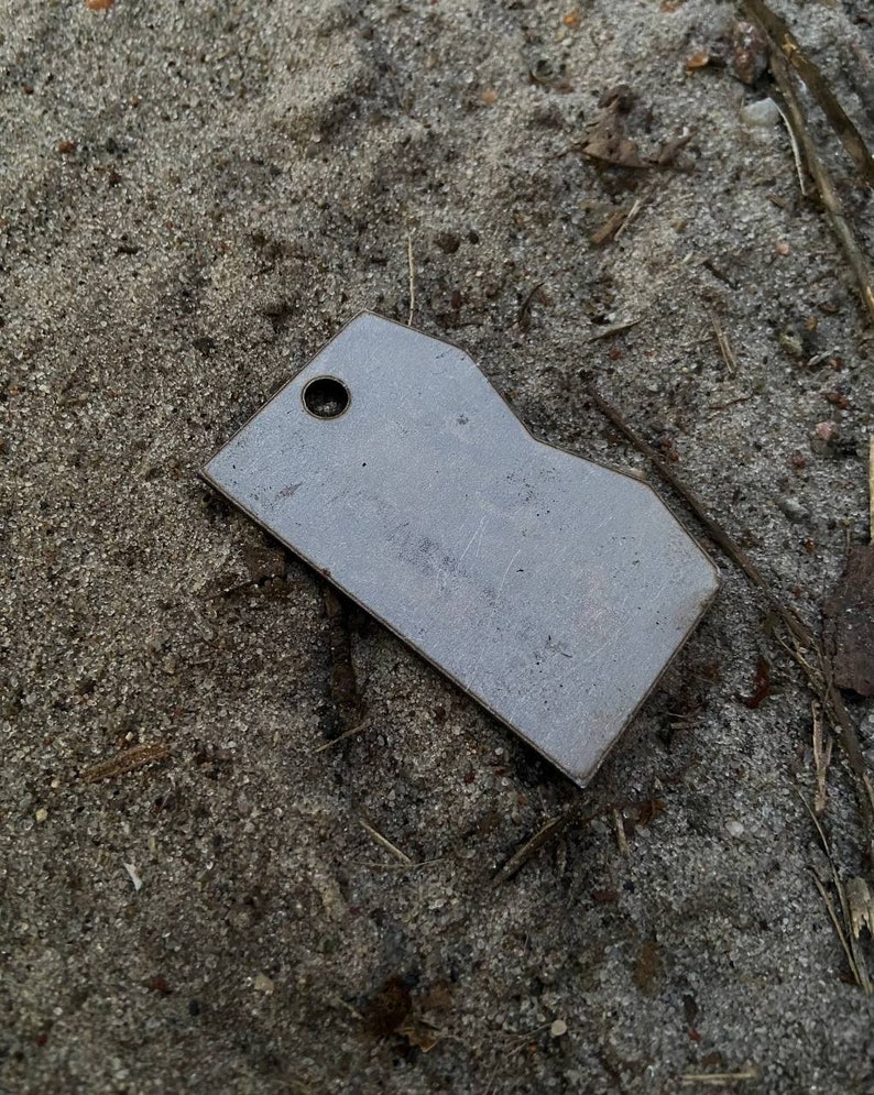 May include: A silver metal tag with a rounded edge and a hole in the top. The tag is lying on a bed of sand.