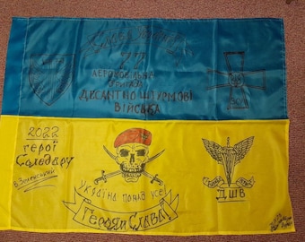 May include: A yellow and blue Ukrainian flag with handwritten text and symbols. The text includes "2022", "Heroi Soleadaru", "V. Zelenskiy", "77 Aeromobilna Brigada", "Desantno Shturmovi Viyska", "Ukraina ponad vse", "Geroyam Slava!" and "DShB". The symbols include a skull and crossbones, a winged sword, and a cross with the Ukrainian trident.