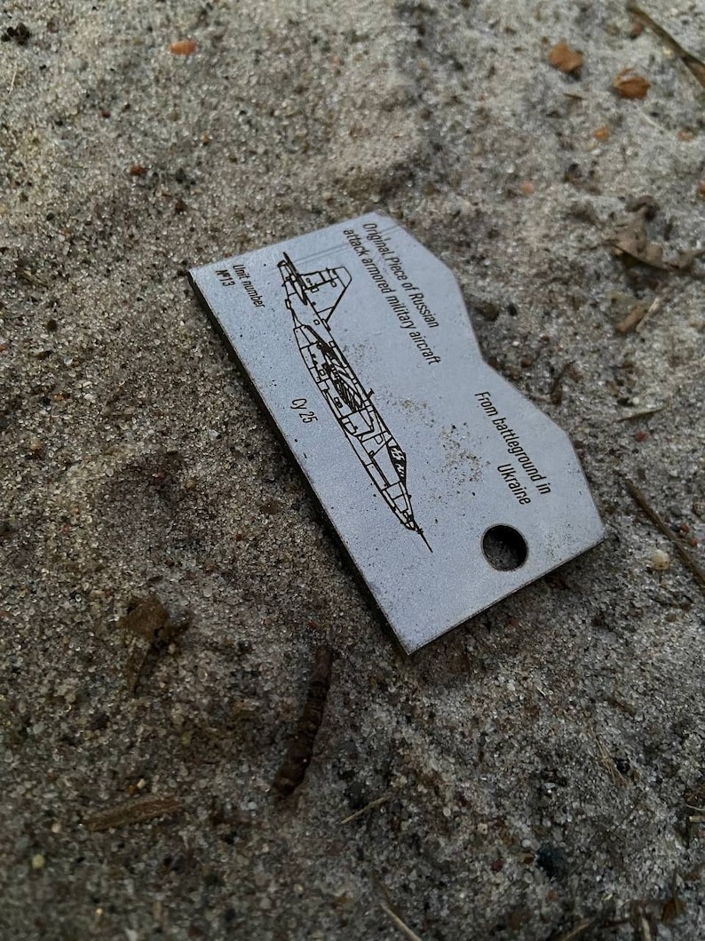 May include: A silver metal tag with a black and white illustration of a Russian attack aircraft. The tag has the text "Original Piece of Russian attack armored military aircraft" and "From battleground in Ukraine". The tag also has the text "Unit number M13" and "Cy 25".