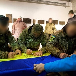 Op de afbeelding: Een groep mensen die een gele en blauwe vlag ondertekenen met zwarte stiften. De vlag ligt op een tafel. De mensen dragen camouflagekleding en één persoon draagt een roze jas.