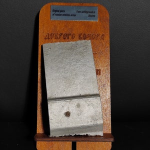 Könnte beinhalten: Ein Stück russischer Fahrzeugpanzerung, das auf einem Holzständer ausgestellt ist. Der Ständer hat eine silberne Plakette mit der Aufschrift "Originalteil der Panzerung russischer Fahrzeuge" und "Vom Schlachtfeld in der Ukraine". Die Panzerung ist hellgrau und hat eine raue Textur. Der Ständer ist dunkelbraun und hat eine glatte Textur.