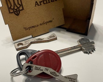 May include: A small wooden box with the words "Ukraine Artifact" and the Ukrainian coat of arms engraved on the lid. The box is open and a small metal key with a piece of a Russian tank attached to it is visible. The key is engraved with the words "Piece of Russian tank T90" and "From Battleground in Ukraine".
