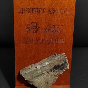 Könnte beinhalten: Holzdisplay mit einem Stück russischer Fahrzeugpanzerung aus der Ukraine. Das Display enthält den Text "Originalteil der russischen Fahrzeugpanzerung" und "Vom Schlachtfeld in der Ukraine."