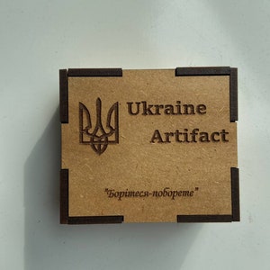 May include: A small wooden box with a brown background and black trim. The box has a Ukrainian trident symbol and the text "Ukraine Artifact" printed on the front. The text "Боринеся-поборете" is printed on the bottom of the box.