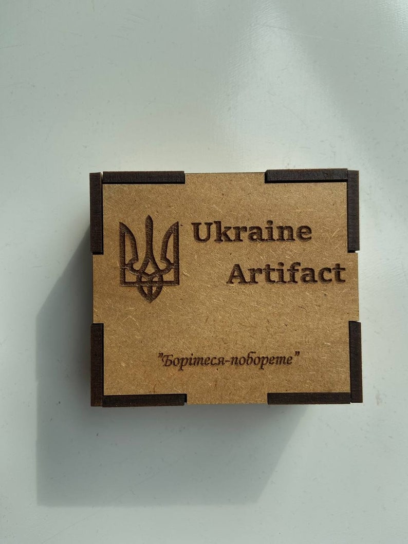 May include: A small wooden box with a brown background and black trim. The box has a Ukrainian trident symbol and the text "Ukraine Artifact" printed on the front. The text "Боринеся-поборете" is printed on the bottom of the box.