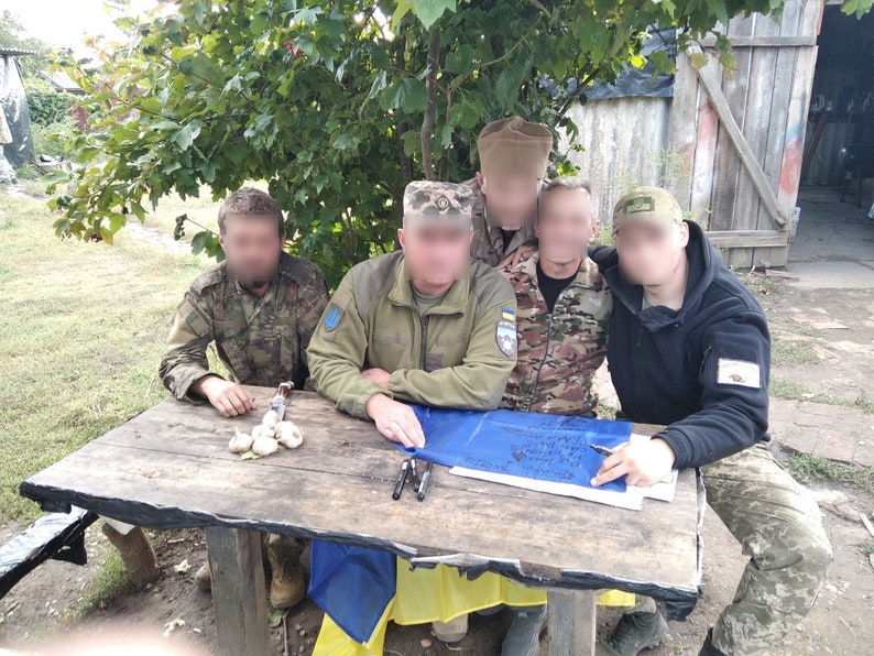May include: A group of five people wearing military uniforms are gathered around a wooden table. They are looking at a blue and yellow flag with a handwritten message on it. The table has a few white objects on it.