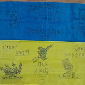 Op de afbeelding: Een blauw en gele vlag met handgetekende illustraties en tekst. De bovenste helft is blauw, de onderste geel. De vlag toont een wapenschild, een kruis, een straaljager en raketwerpers. Tekst bevat Oekraïense zinnen.