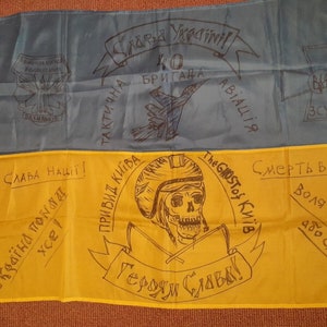 May include: A yellow and blue Ukrainian flag with handwritten messages and drawings. The flag features a skull and crossbones with the words "The Ghost of Kyiv" and "Glory to the Heroes!" The flag also has a drawing of a jet fighter and the words "40th Brigade, Tactical Aviation".