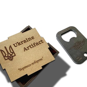 Może przedstawiać: Drewniane pudełko z pokrywką, na której widnieje napis "Ukraine Artifact" oraz herb Ukrainy. Pokrywka zawiera również tekst "Боримесь-побореме". Obok pudełka znajduje się metalowy otwieracz do butelek z wzorem czołgu.