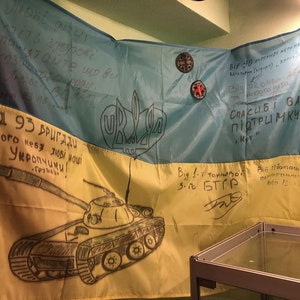 Puede incluir: Una bandera ucraniana amarilla y azul con mensajes escritos a mano en ucraniano. La bandera está colgada sobre una mesa y tiene un dibujo de un tanque en la sección amarilla. El texto en la bandera dice "Від 93 бригади Мирного неба любі наші Укропчики!" y "Від 1-ї тонкової 3-10 БТГР".