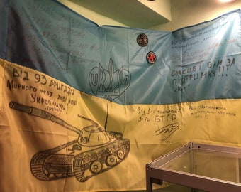 May include: A yellow and blue Ukrainian flag with handwritten messages in Ukrainian. The flag is draped over a table and has a drawing of a tank on the yellow section. The text on the flag reads "Від 93 бригади Мирного неба любі наші Укропчики!" and "Від 1-ї тонкової 3-10 БТГР".