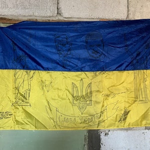 May include: A blue and yellow Ukrainian flag with a trident symbol and the words "Слава Україні!" written in black. The flag is decorated with drawings of people and buildings.