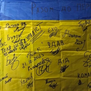 Op de afbeelding: Een gele en blauwe vlag met de woorden "РАЗОМ ДО ПЕРЕМОГИ" in zwarte stift geschreven. De vlag is bedekt met handtekeningen en data, waaronder "21.12.2023" en "Харків".