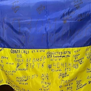 Può includere: Una bandiera ucraina gialla e blu con messaggi scritti a mano con un pennarello nero. I messaggi includono numeri di unità, date e luoghi. La bandiera è firmata da soldati.