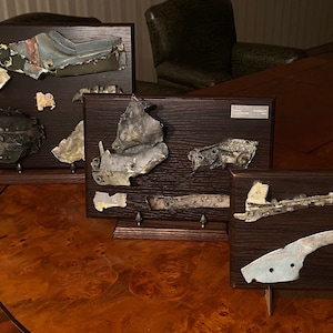 May include: Four framed displays of aircraft debris, each with a dark wood frame and a black background. The debris is made of metal and is painted in various shades of grey, blue, and green. The frames are mounted on a wooden table.