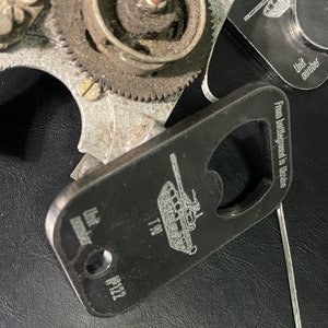 Puede incluir: Un abridor de botellas de metal negro con un diseño de tanque y el texto "From battlefield in Ukraine" y "T-90". El abridor de botellas tiene un agujero en la parte superior y el texto "Unit number" está grabado en el lateral.