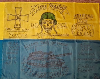 May include: A Ukrainian flag with handwritten messages in black marker. The top half of the flag is yellow with a black cross and the text "Слава Україні" (Glory to Ukraine), "ЗСУ" (Armed Forces of Ukraine), "Збройні сили України" (Armed Forces of Ukraine), "ESTD 2022", "ІнтернаціонАЛЬНИЙ ЛЕГІОН" (International Legion), "INTERATIONAL LEGION", "DEFENCE of UKRAINE". The bottom half of the flag is blue with the text "WAR 2022", "DEFENCE", "Київ" (Kyiv), "KYIV", "Abrams tanks for Ukraine", "Війна 2022" (War 2022), "Оборона" (Defense), "Сіверодонецьк" (Severodonetsk), "Соледар", "Бахмут". A black and white drawing of a tank is in the center of the blue half of the flag.