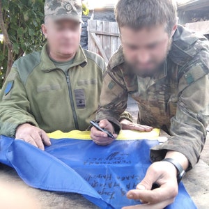 May include: Two people in military uniforms are signing a blue and yellow Ukrainian flag with black markers. The flag is laid out on a wooden table.